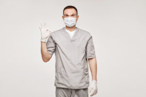 The image features a male healthcare professional in a grey medical uniform, wearing a surgical mask and gloves, giving a reassuring gesture with his hand raised in an "OK" sign, signaling readiness and safety in a clinical or hospital setting.