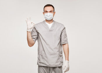 The image features a male healthcare professional in a grey medical uniform, wearing a surgical mask and gloves, giving a reassuring gesture with his hand raised in an "OK" sign, signaling readiness and safety in a clinical or hospital setting.