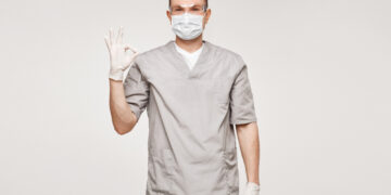 The image features a male healthcare professional in a grey medical uniform, wearing a surgical mask and gloves, giving a reassuring gesture with his hand raised in an "OK" sign, signaling readiness and safety in a clinical or hospital setting.