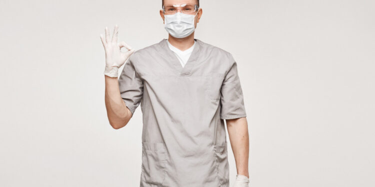 The image features a male healthcare professional in a grey medical uniform, wearing a surgical mask and gloves, giving a reassuring gesture with his hand raised in an "OK" sign, signaling readiness and safety in a clinical or hospital setting.